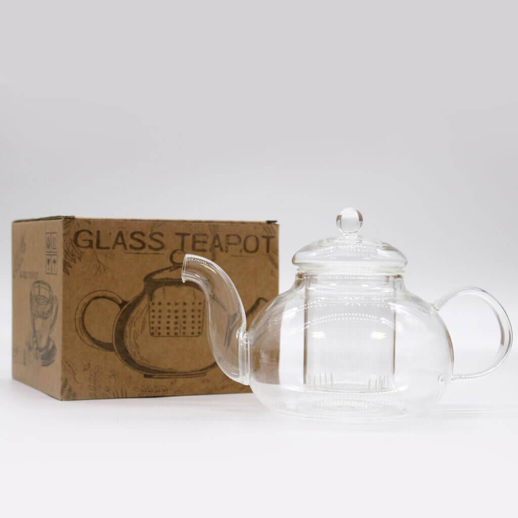 A clear glass teapot with a lid is placed next to a brown cardboard box labeled "GLASS TEAPOT" on a plain white background.