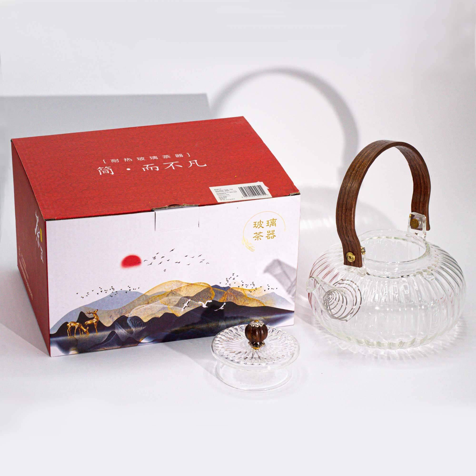 A clear glass teapot with a wooden handle and lid sits next to a red and white box featuring mountain and animal illustrations.