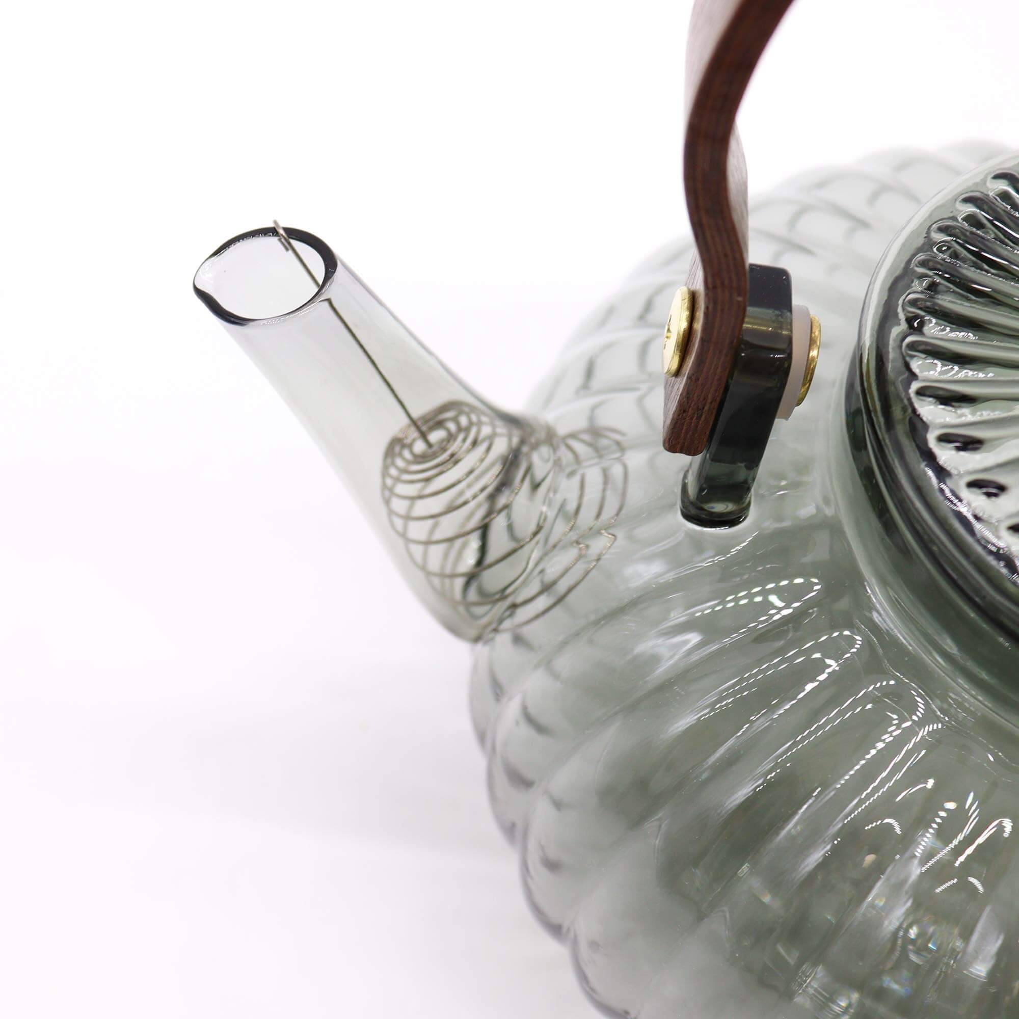 Close-up of a transparent glass teapot with a ribbed design, wooden handle, and a visible metal filter inside the spout.