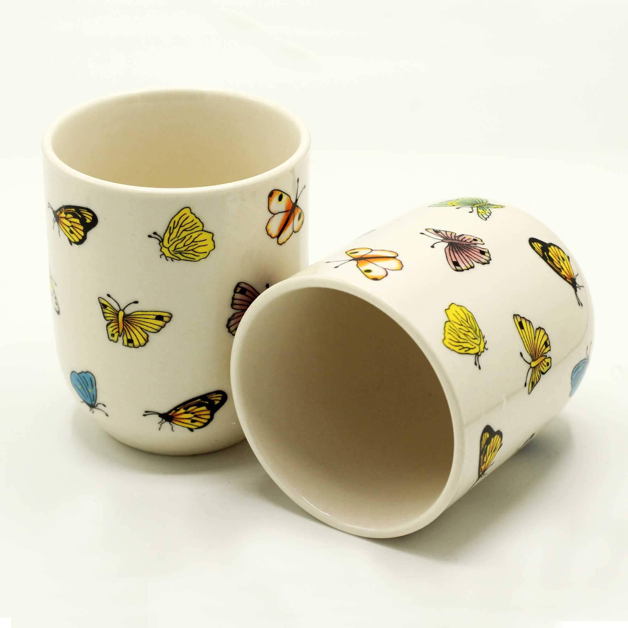 Two white ceramic cups with colorful butterfly and leaf patterns; one cup stands upright while the other lies on its side.