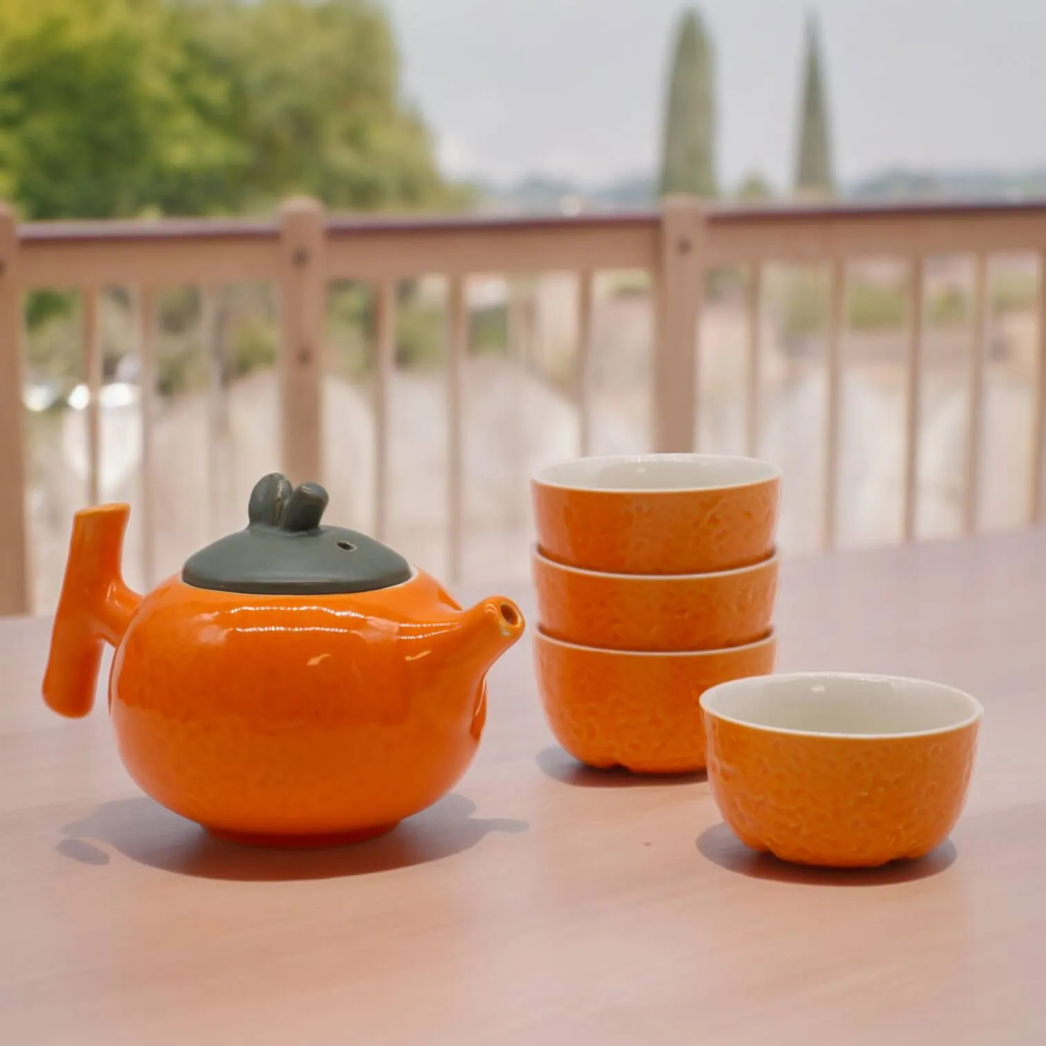 An orange teapot with a lid and three matching teacups are arranged on a light wooden table, with a blurred outdoor background.