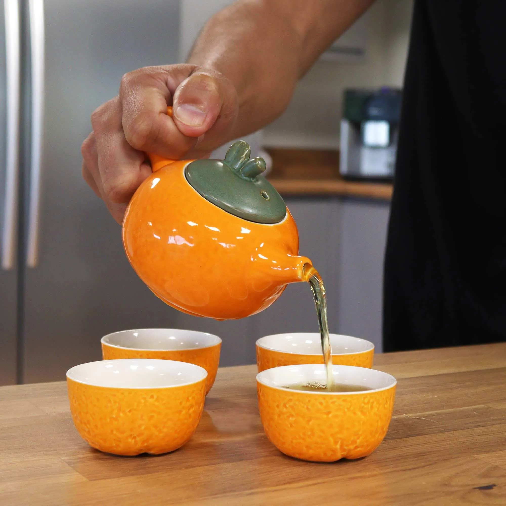A hand pours tea from an orange teapot into one of four matching orange teacups on a wooden countertop in a kitchen.