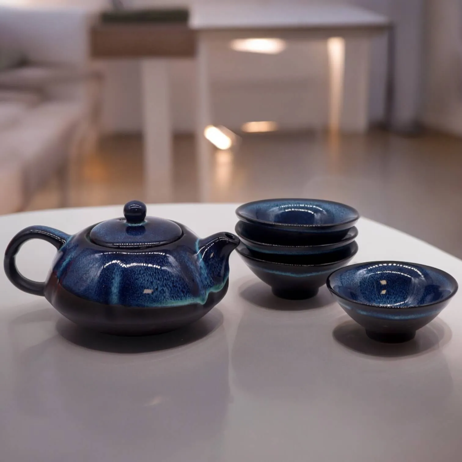 A dark blue ceramic teapot is placed on a white table next to four matching teacups, three of which are stacked. The background is softly lit.