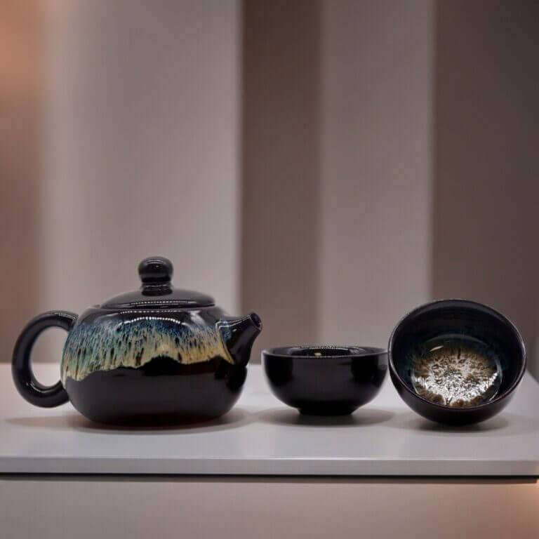 A black ceramic teapot with a textured glaze sits next to two matching teacups on a white surface against a softly blurred background.
