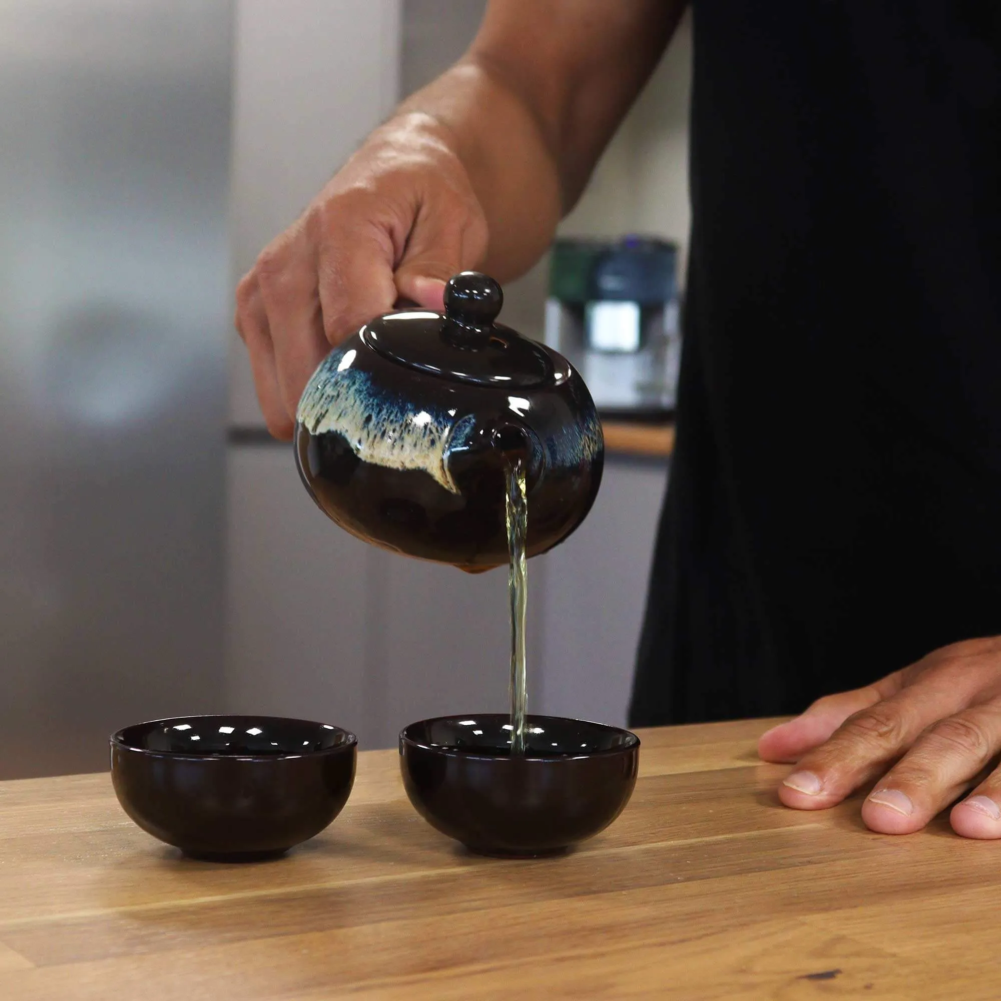 A person pours tea from a ceramic teapot into one of two black teacups on a wooden table.