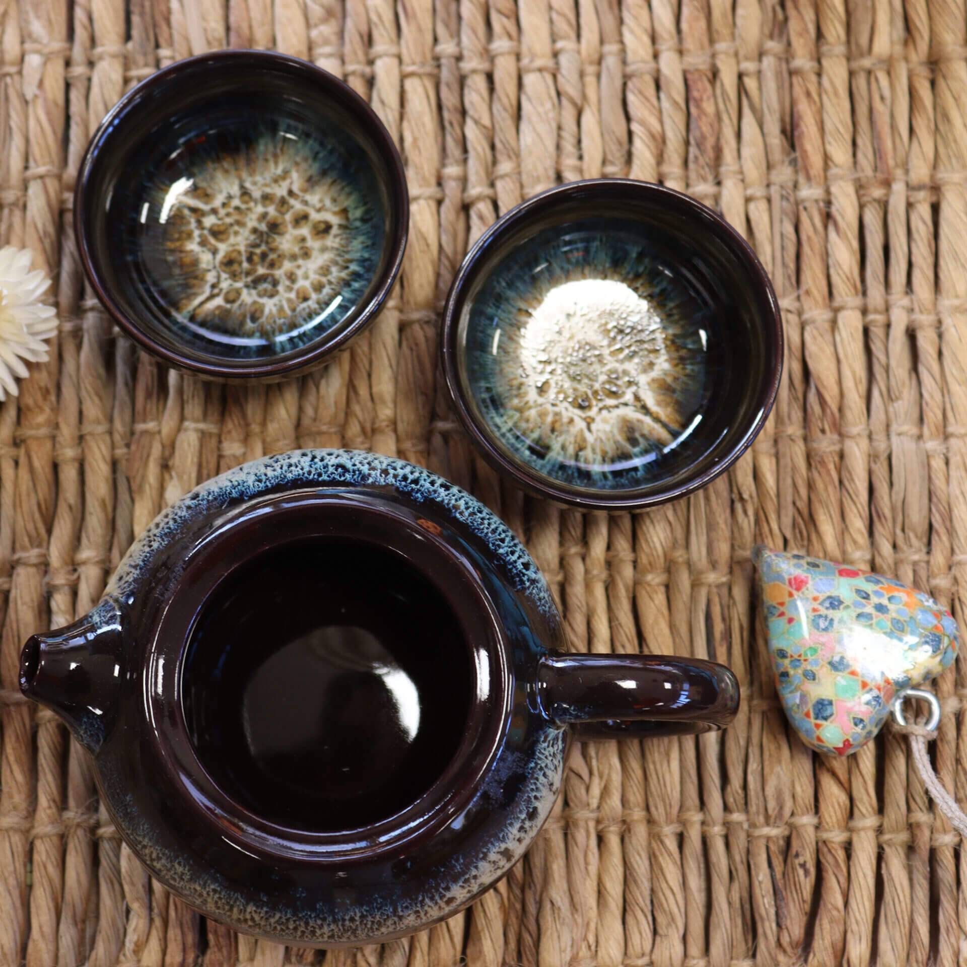 A dark ceramic teapot with a matching pair of cups and a floral-patterned ceramic heart on a woven mat.