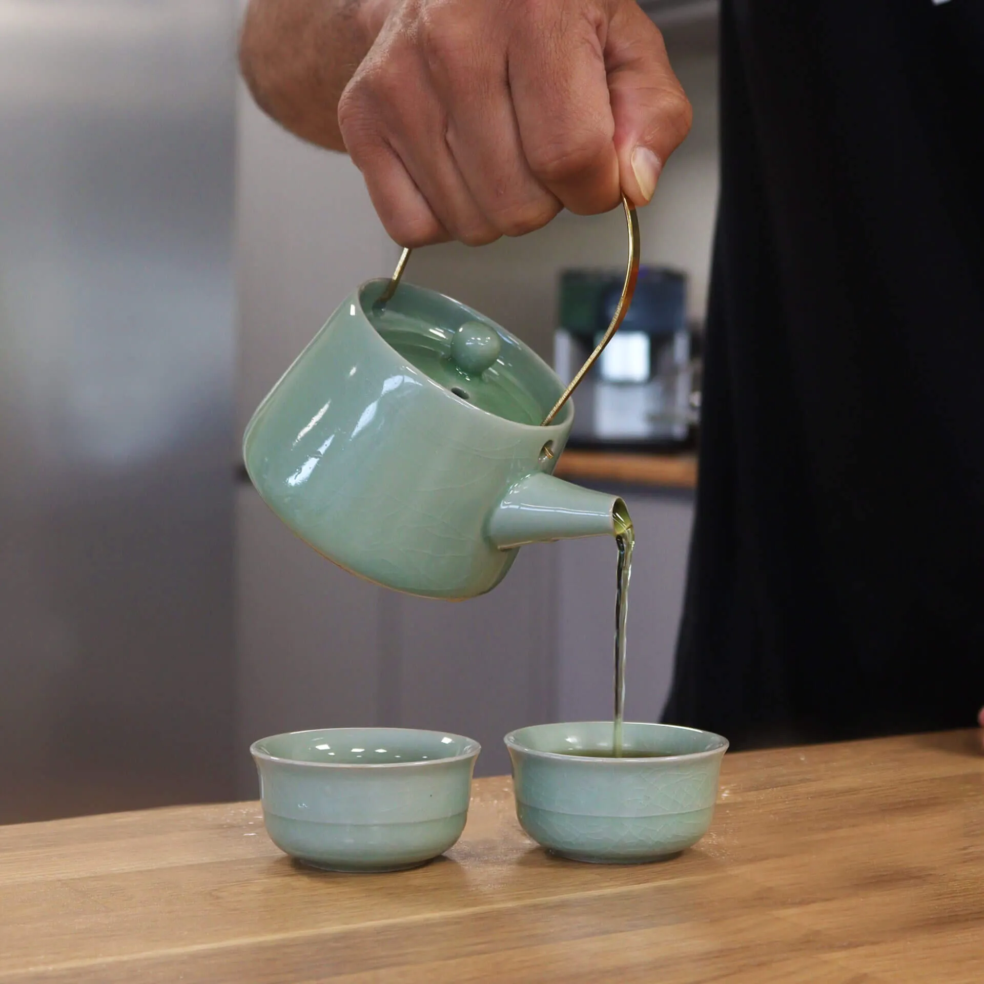 A hand pours tea from a green teapot into one of two matching cups on a wooden surface.