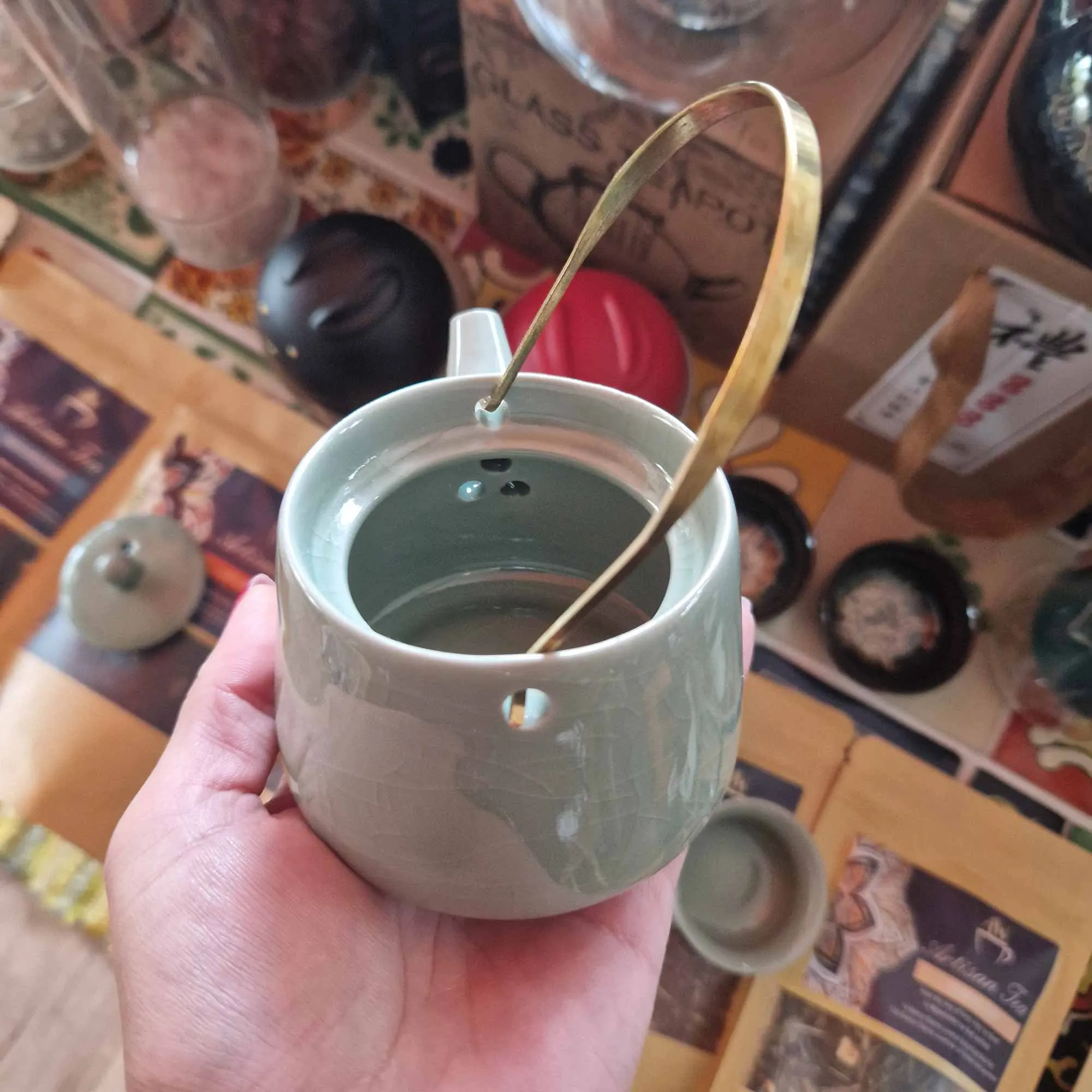 A hand holds a small ceramic teapot with a brass handle, showing its open top and lid, surrounded by tea sets and packaging on a wooden surface.
