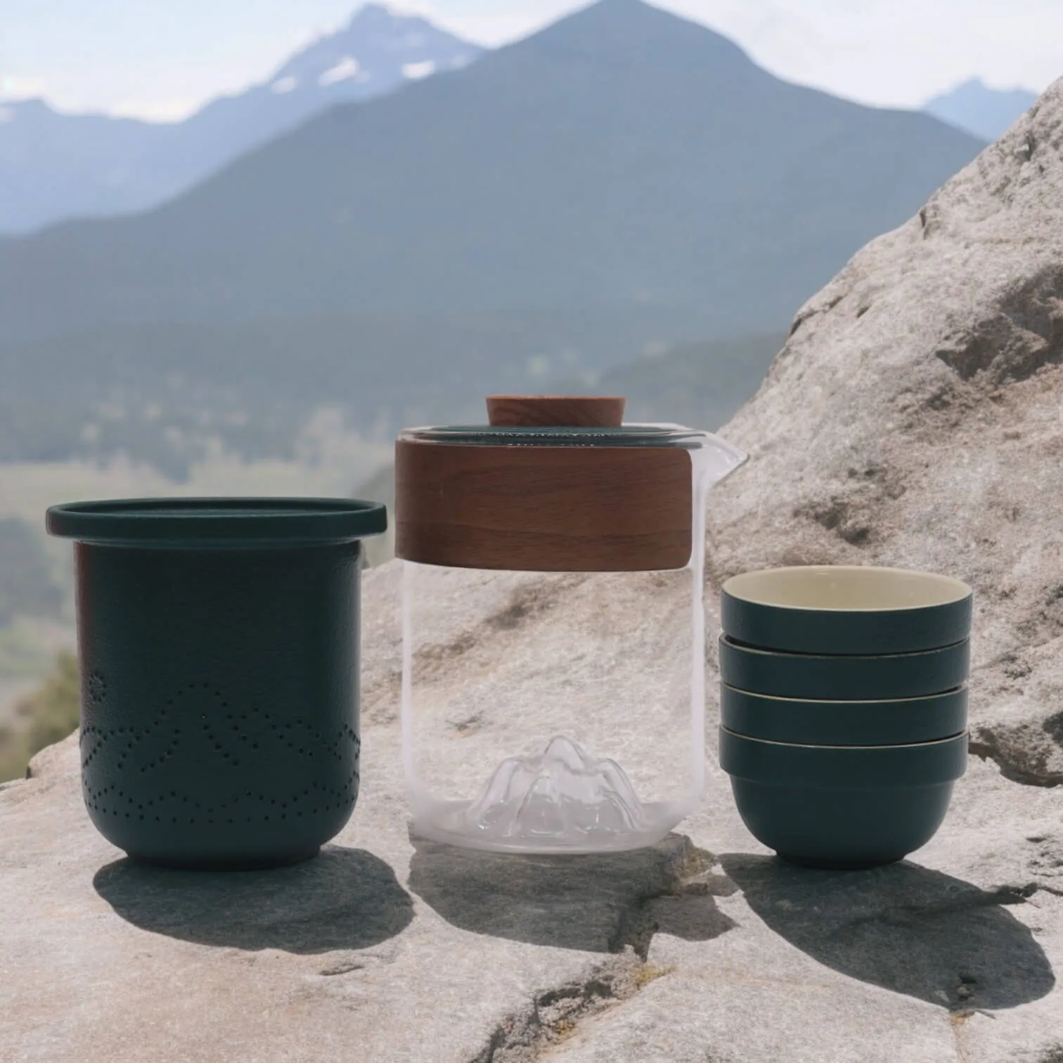 A glass pitcher with a wooden lid, a dark green cup, and a stack of three matching cups are placed on a rock with mountains in the background.