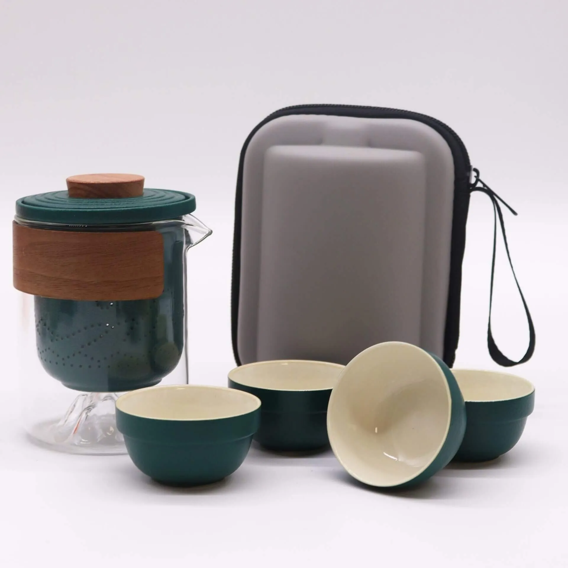 A portable tea set with a glass teapot, wooden and green lid, four green and cream cups, and a gray carrying case with a black zipper and strap.