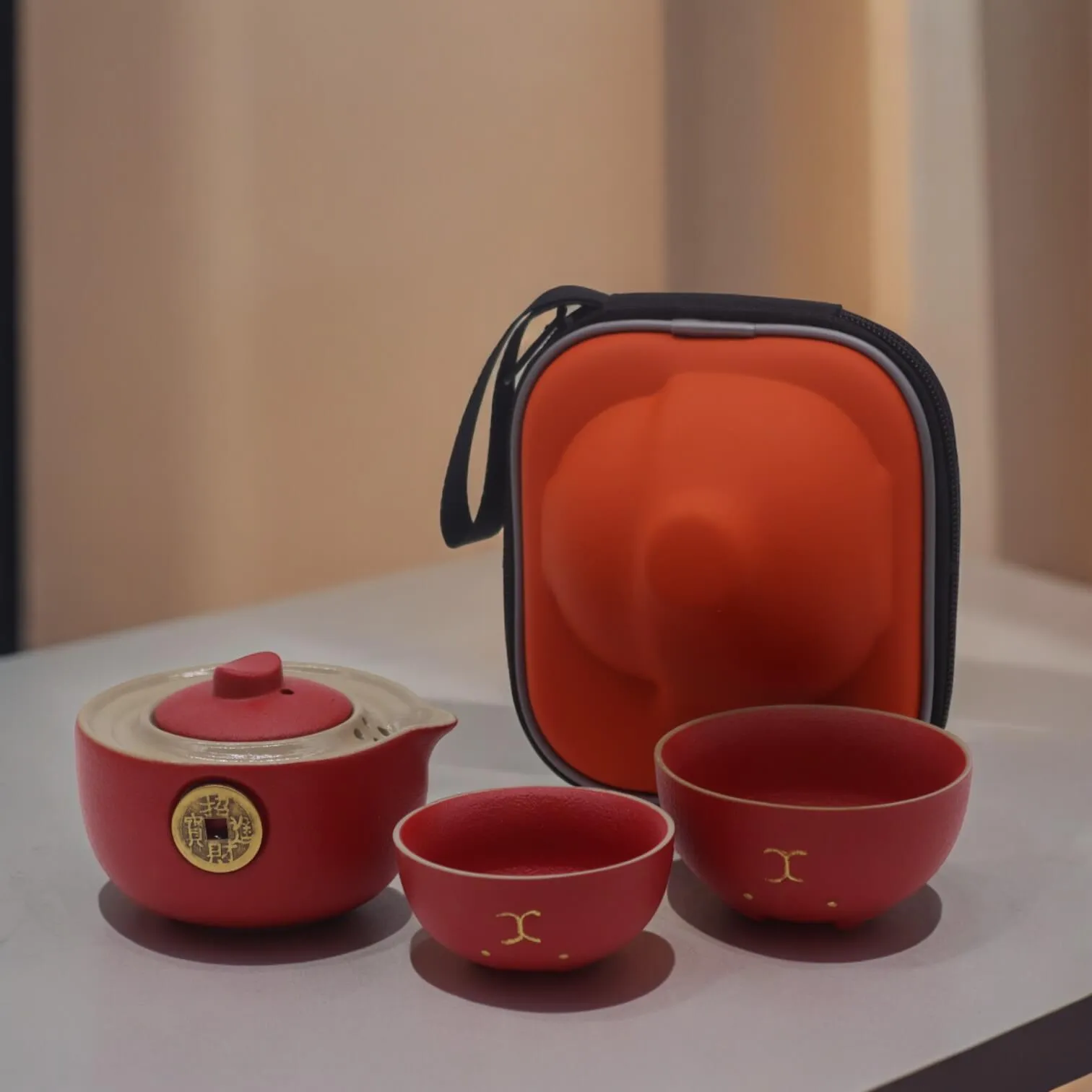 A red and beige teapot with two matching cups is displayed next to a red and black zippered carrying case on a light-colored surface.