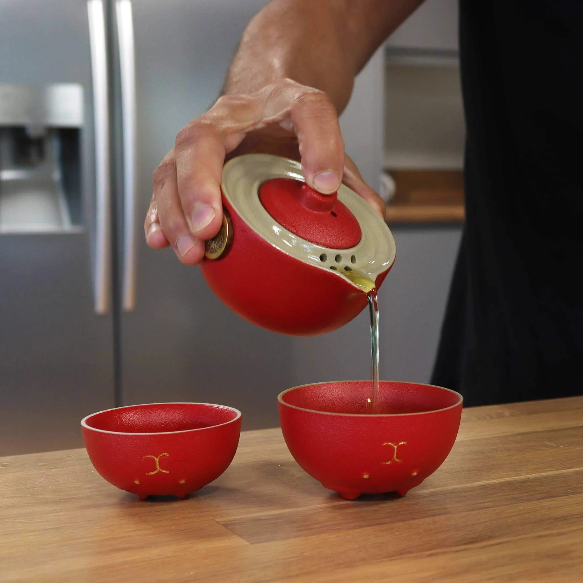 A person pours tea from a red teapot into one of two matching red cups on a wooden countertop in a kitchen.