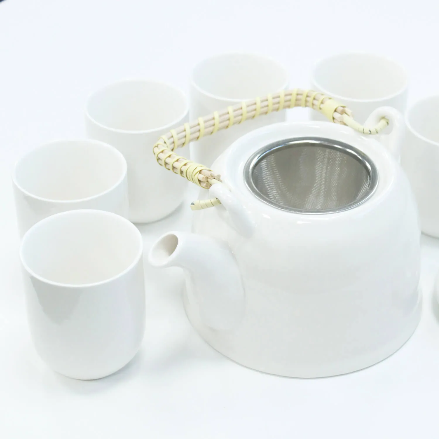 A white teapot with a woven handle and metal strainer is surrounded by six matching white cups on a white surface.