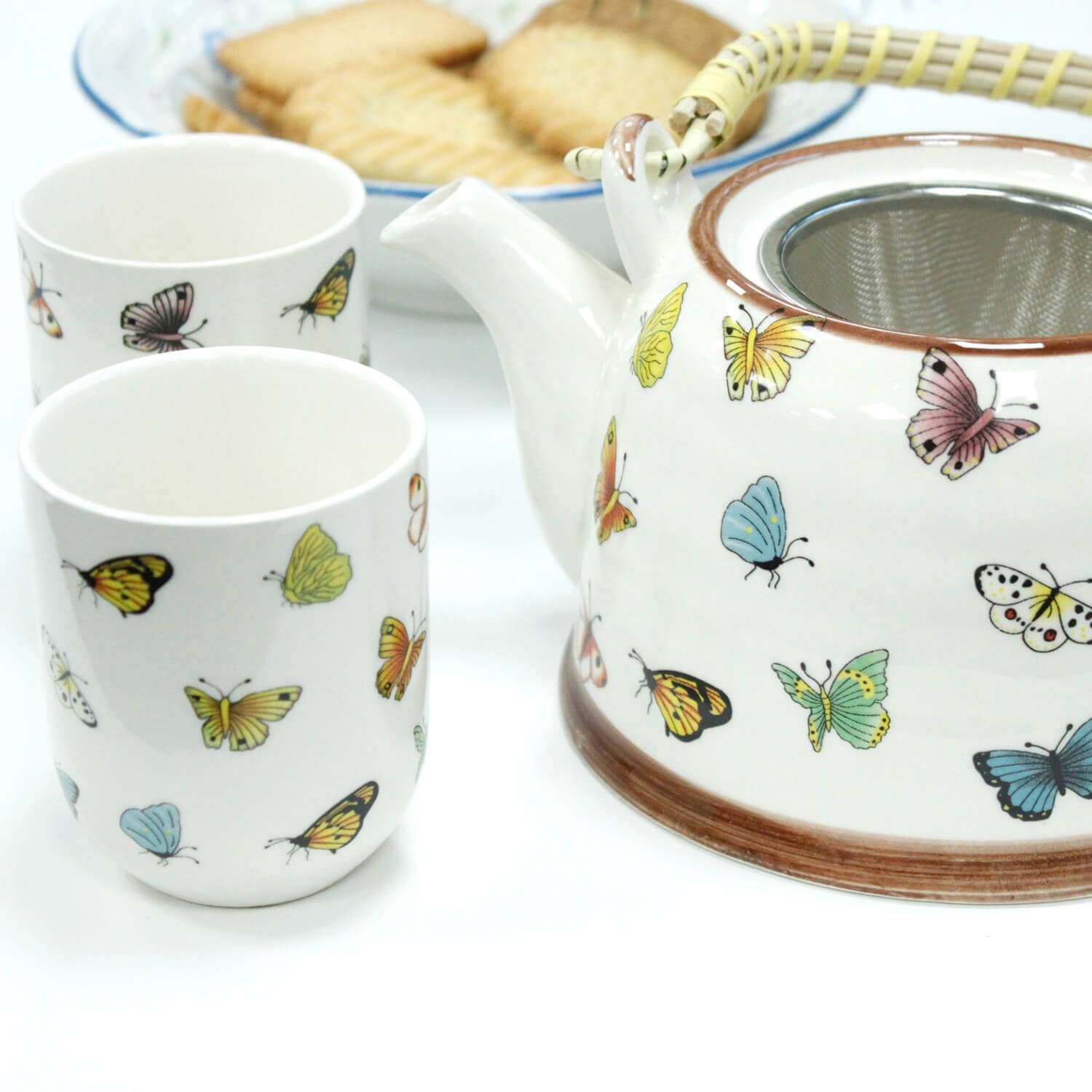 A white teapot and two cups decorated with colorful butterfly designs are placed next to a plate of cookies.