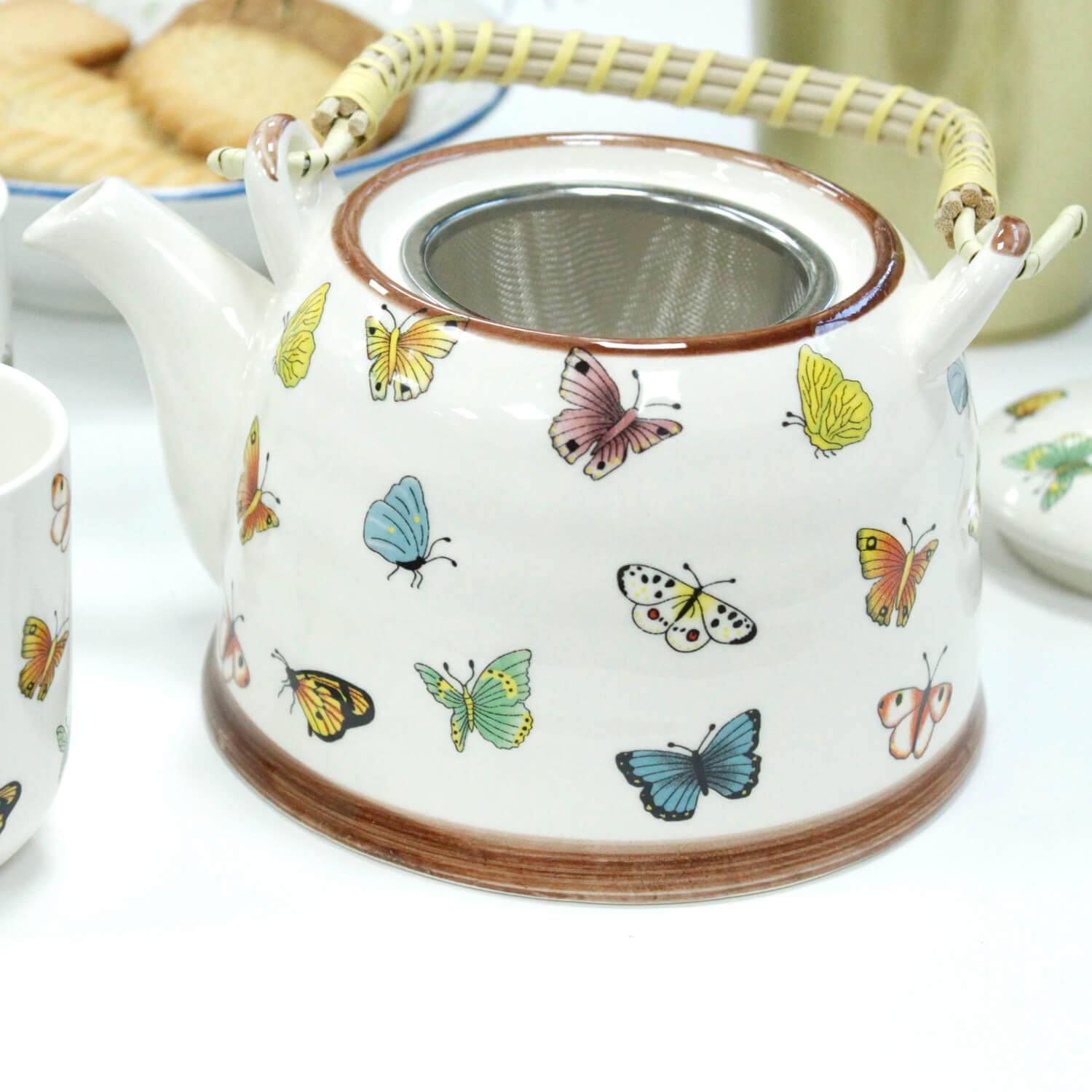 A white ceramic teapot with a bamboo handle is decorated with colorful butterfly illustrations. A metal infuser is visible inside the teapot.