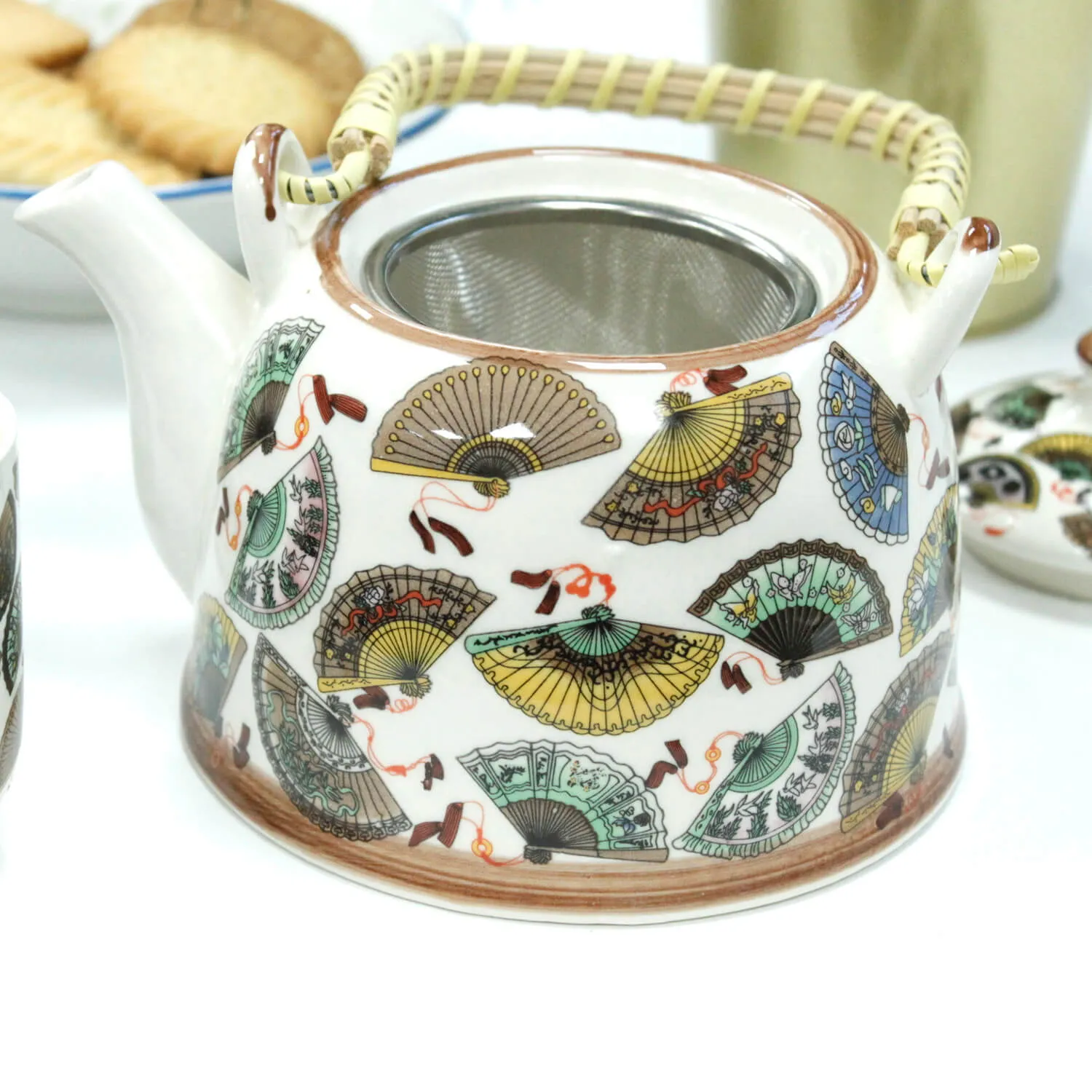 A ceramic teapot with a woven handle, decorated with colorful fan patterns and an integrated metal strainer, sits on a white surface.