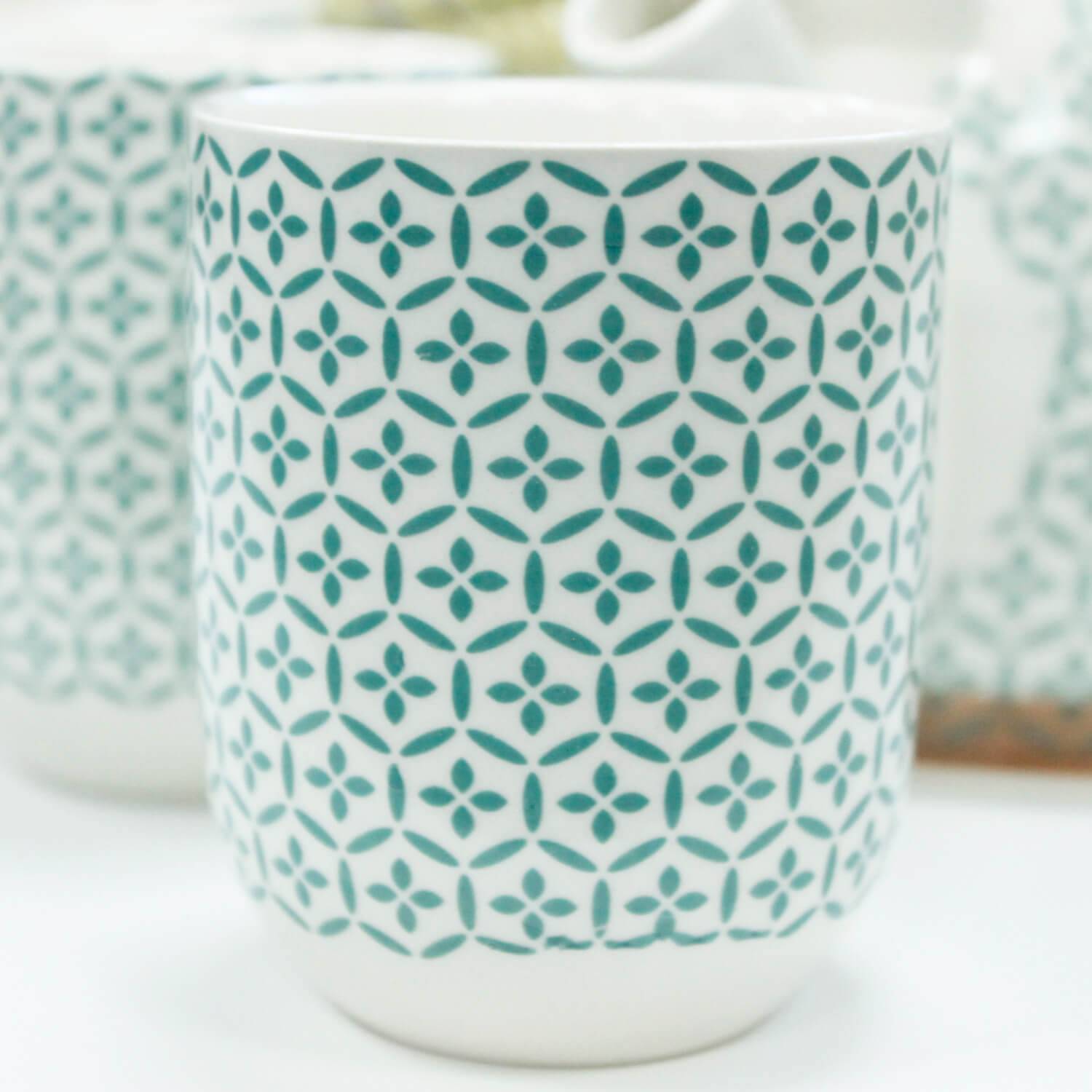 White ceramic cup with a teal geometric pattern consisting of repeating diamond and circle shapes. The cup is on a white surface, with another similar cup slightly blurred in the background.
