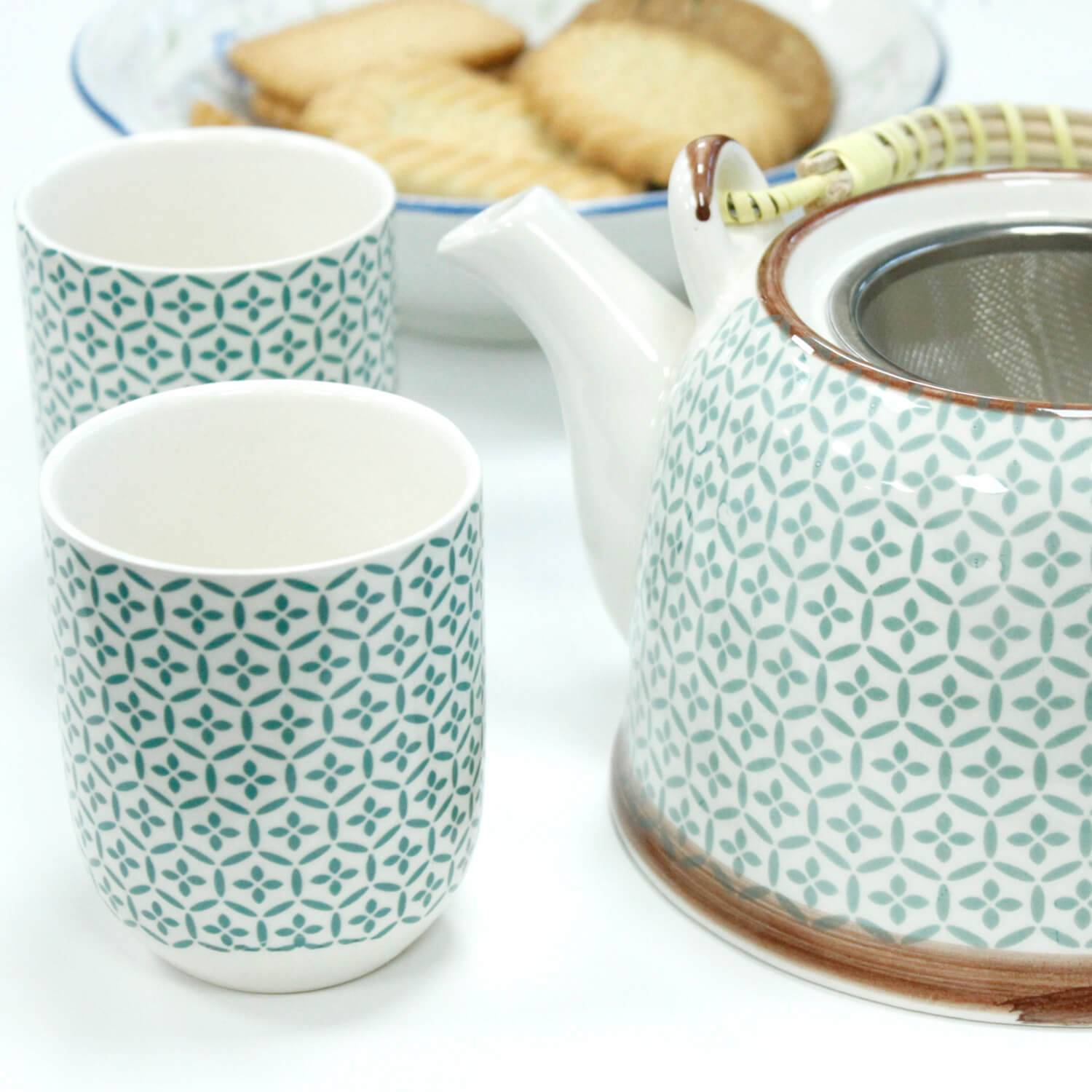 A patterned ceramic teapot with a matching handle is next to two matching teacups; a plate of cookies is in the background.