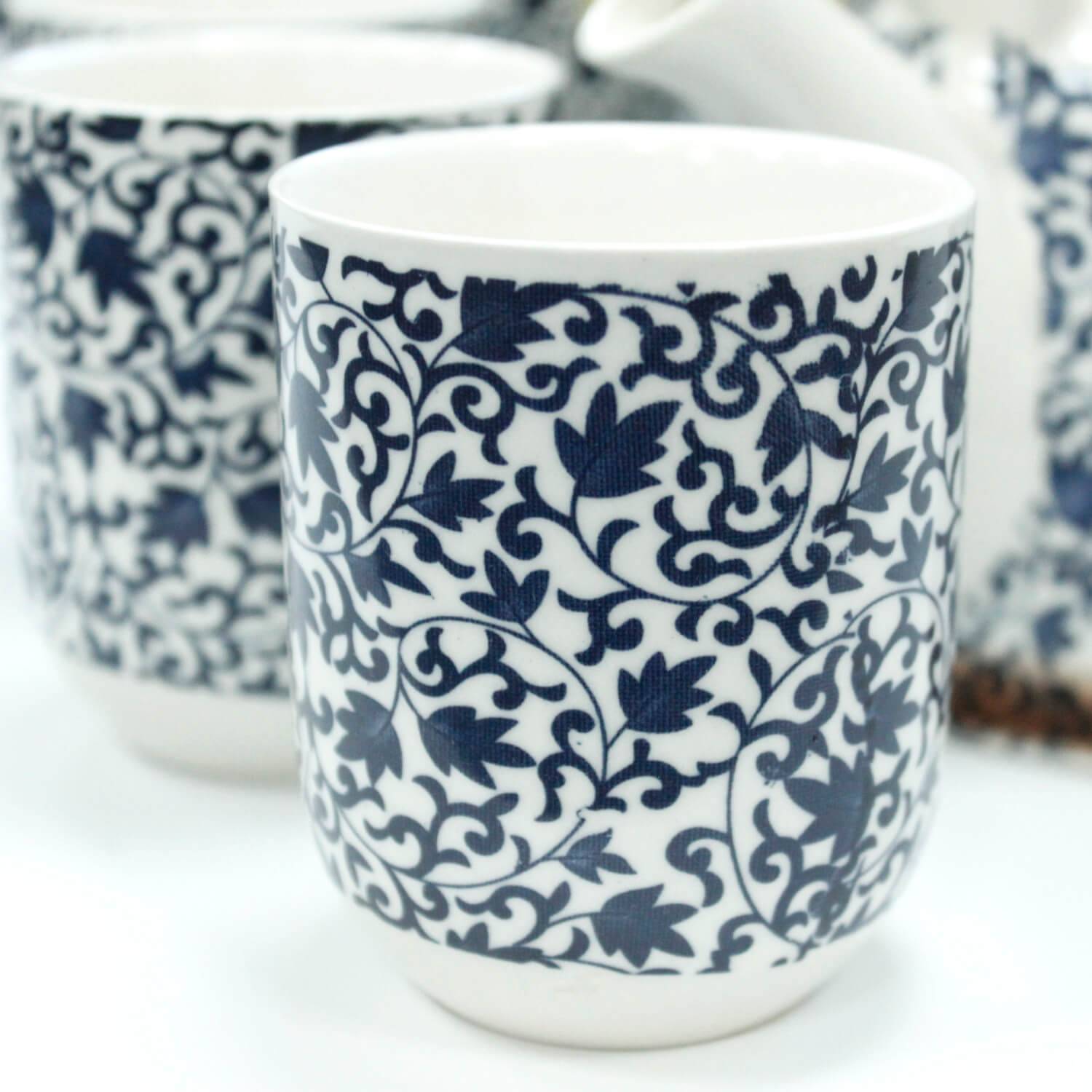 A white ceramic cup featuring an intricate blue floral vine pattern, with parts of matching cups and a teapot visible in the background.