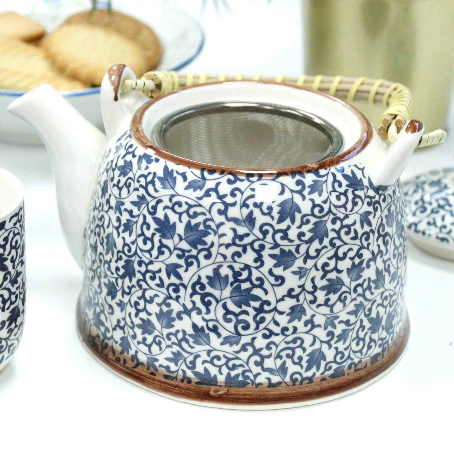 A ceramic teapot with a blue floral pattern and a bamboo handle, shown with a metal strainer inside and some cookies in the background.