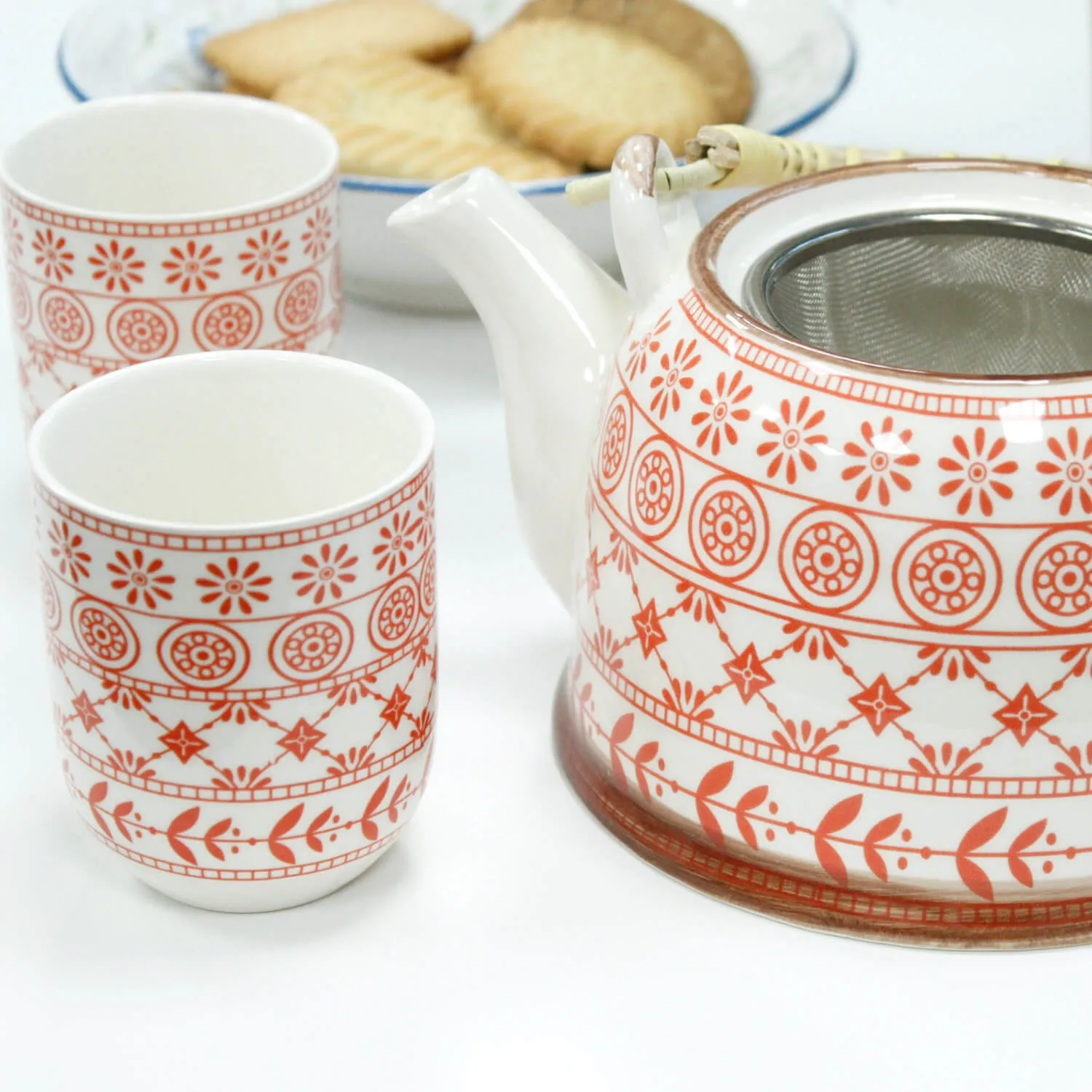 A white teapot and two matching cups with red patterns are placed next to a plate of rectangular biscuits.