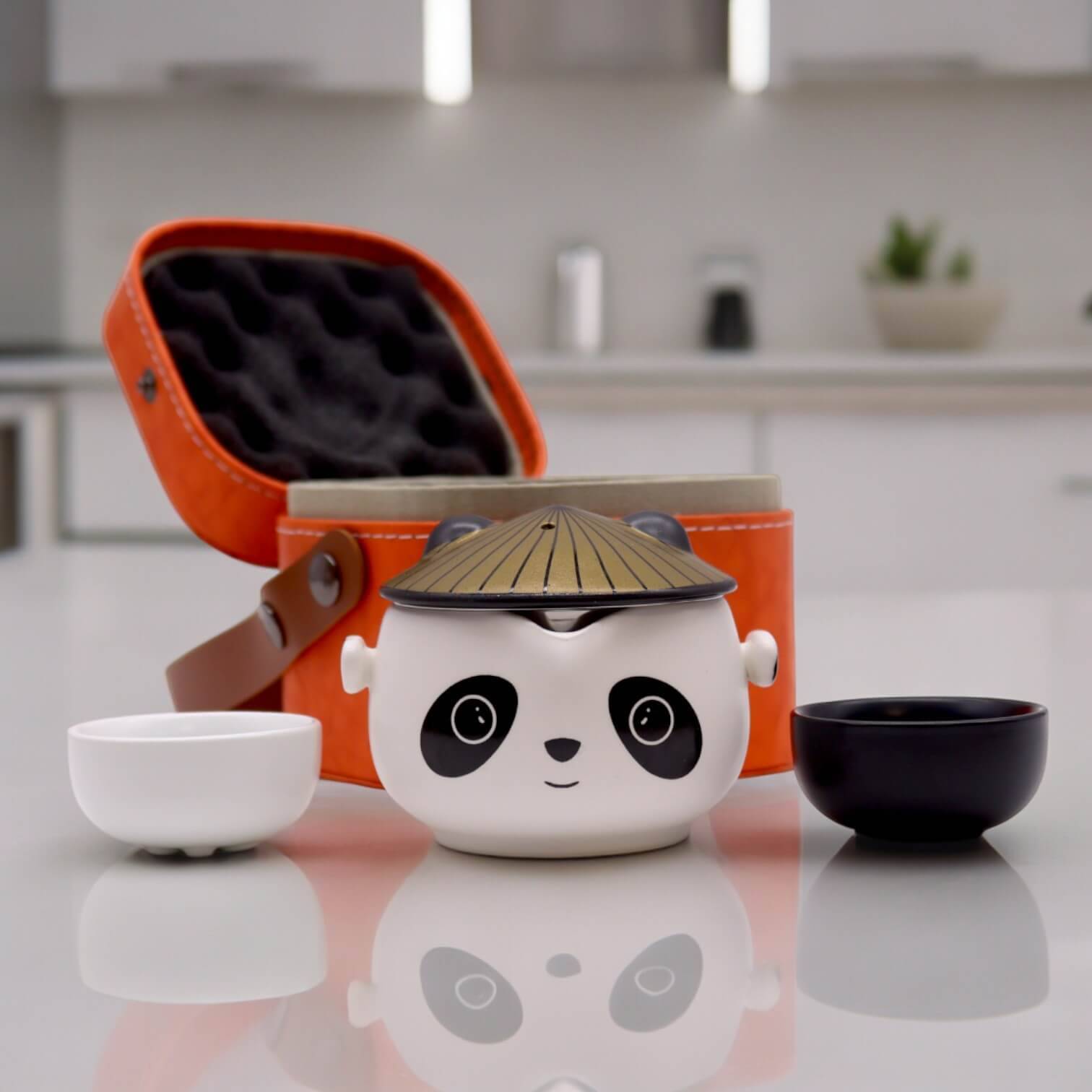 A panda-shaped tea set with two small cups and an orange carrying case is displayed on a kitchen countertop.