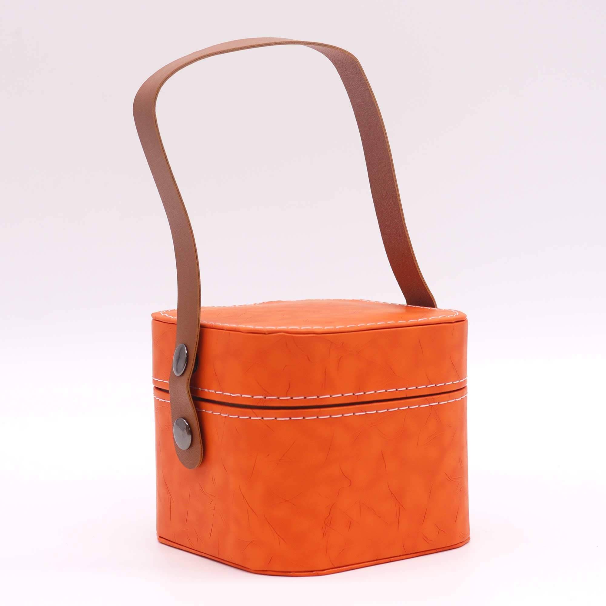 An orange, hexagonal leather container with white stitching and a brown handle attached by two metal studs on each side.