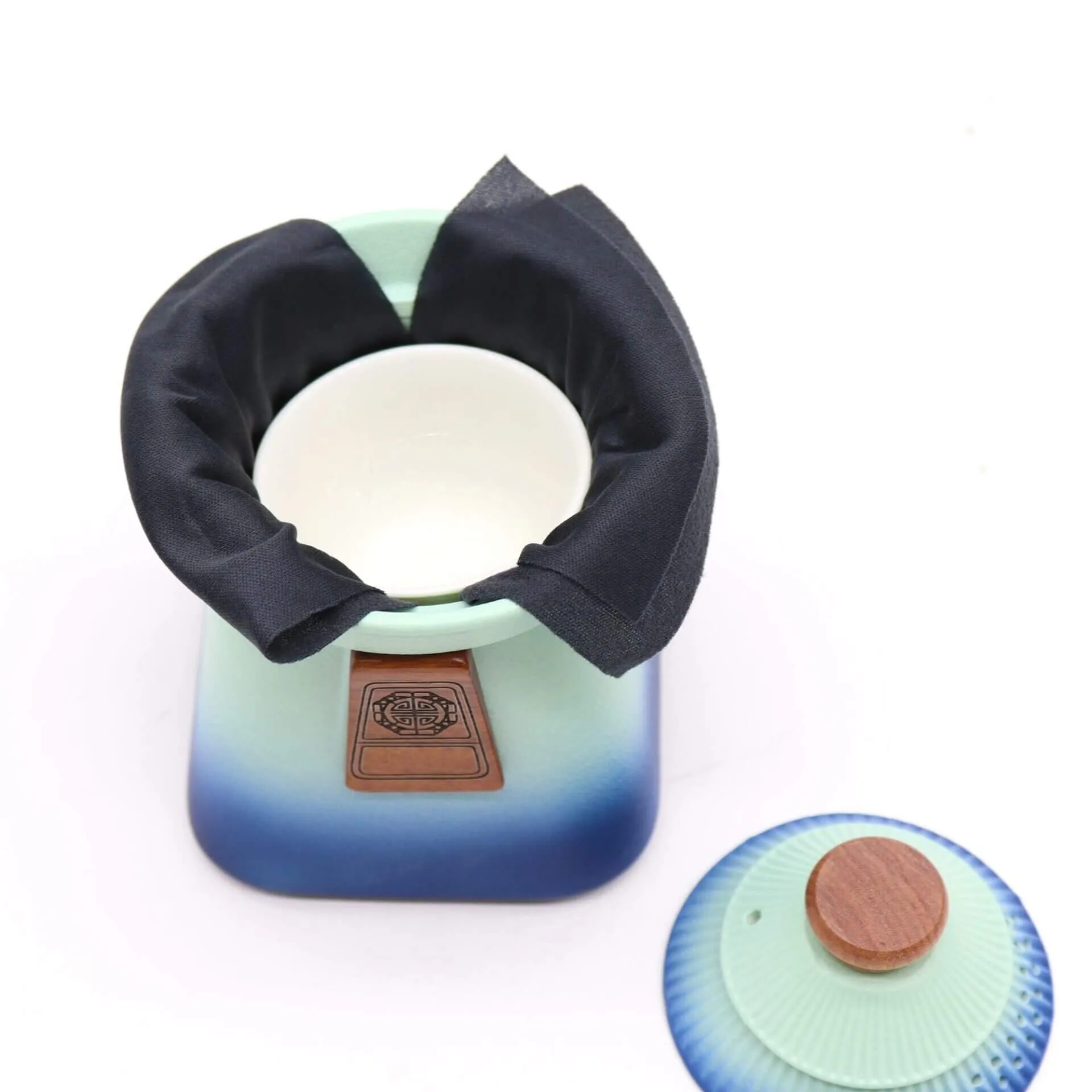 A ceramic container with a cloth covering its opening sits beside a matching blue lid with a wooden knob on a white background.