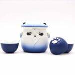 A blue and white panda-shaped teapot with a lid, flanked by two matching teacups, one turned to show a white paw print on its base.