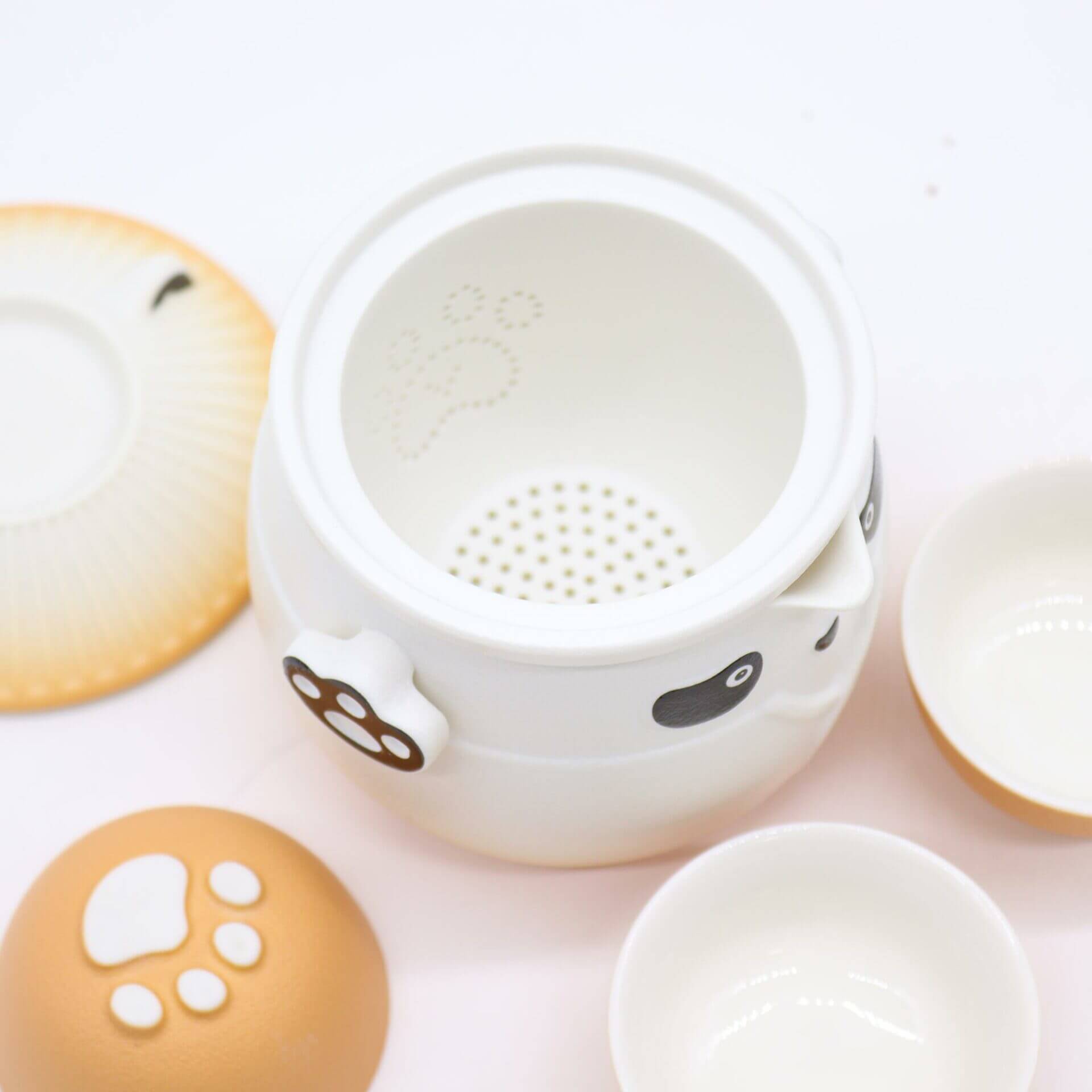 A white ceramic teapot with panda face and paw print designs, accompanied by a matching lid, two small teacups, and a saucer on a white surface.