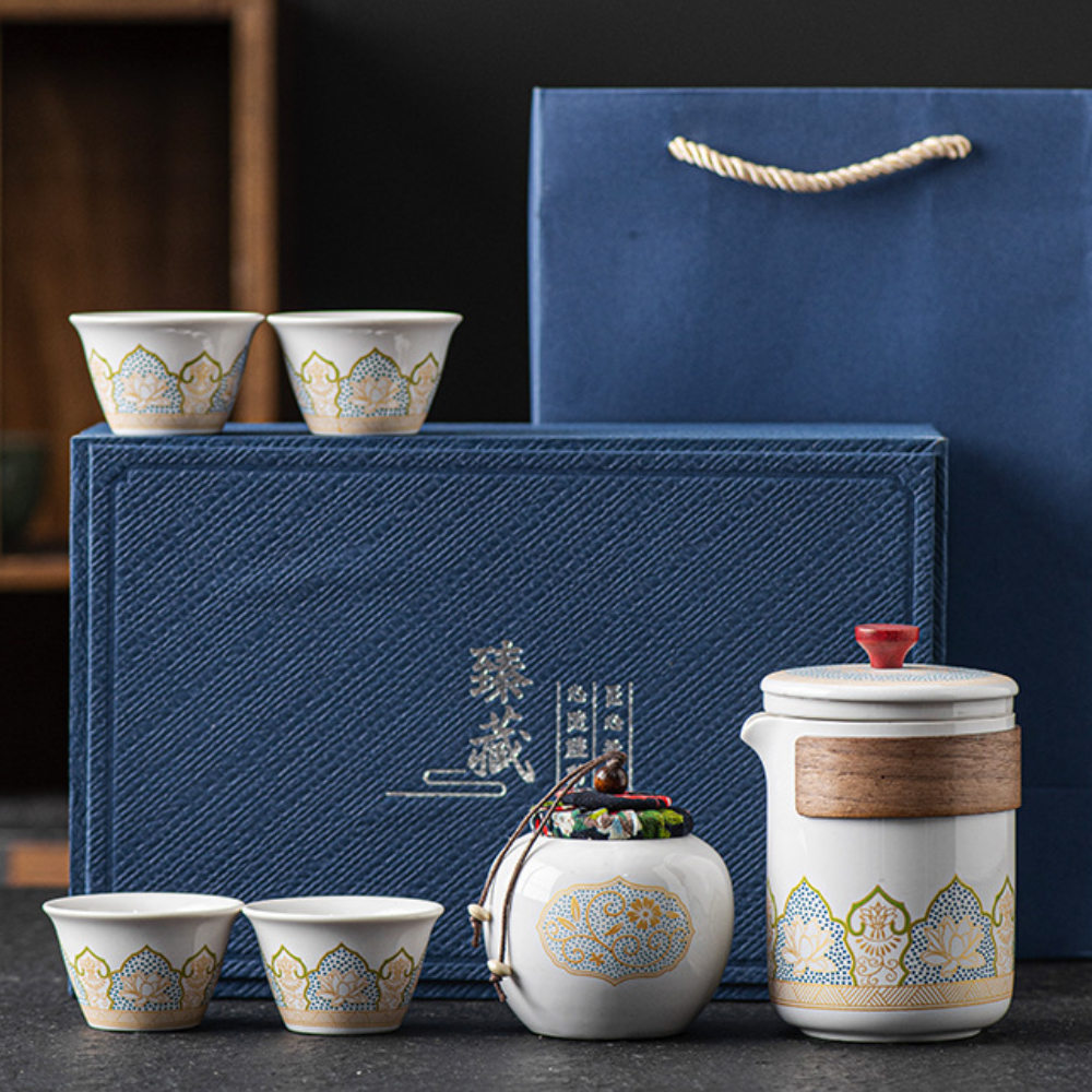 A ceramic tea set with four cups, a lidded teapot, a small jar, a blue textured box, and a blue gift bag are arranged on a dark surface.
