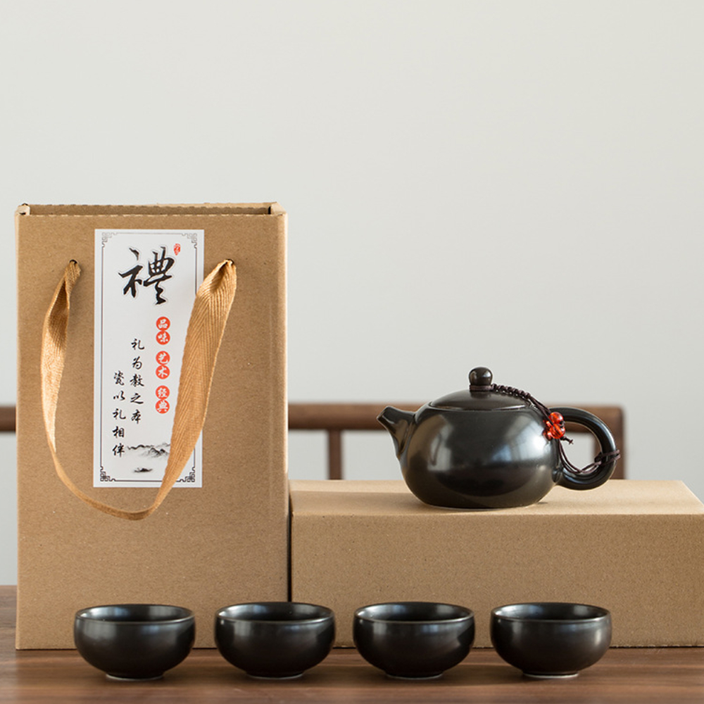 A black ceramic teapot with four matching teacups is displayed beside two brown gift boxes, one with rope handles and Chinese characters on a white label.