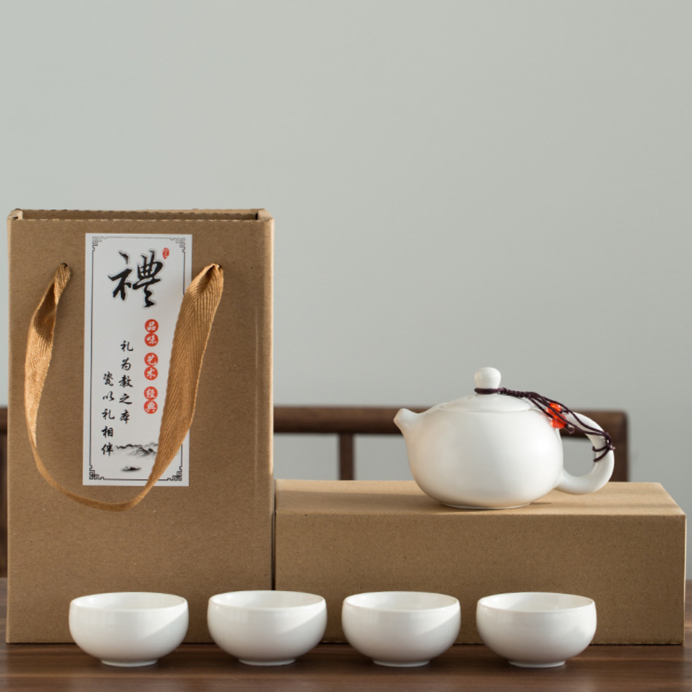 A white ceramic teapot and four matching teacups are displayed in front of a cardboard gift box with Chinese writing and a fabric handle.