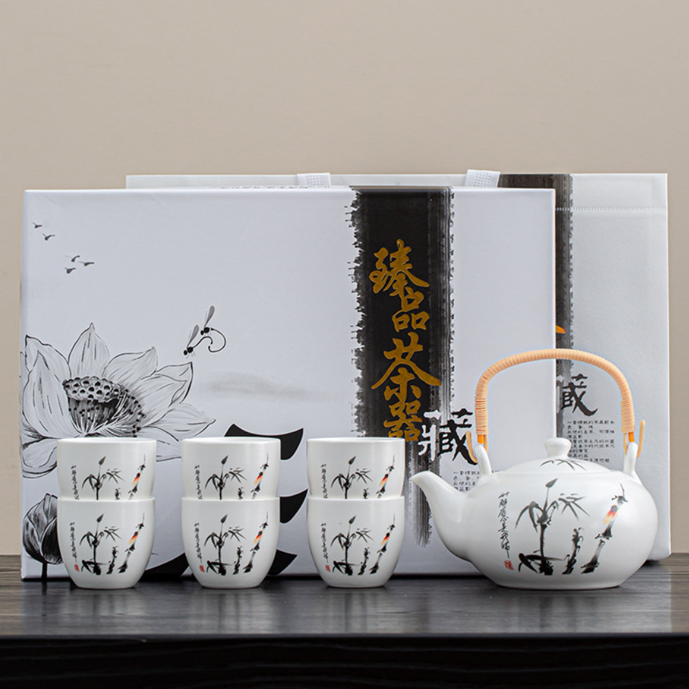 A white porcelain tea set with black and orange designs, including a teapot and six cups, is arranged in front of two decorative boxes and a white bag.