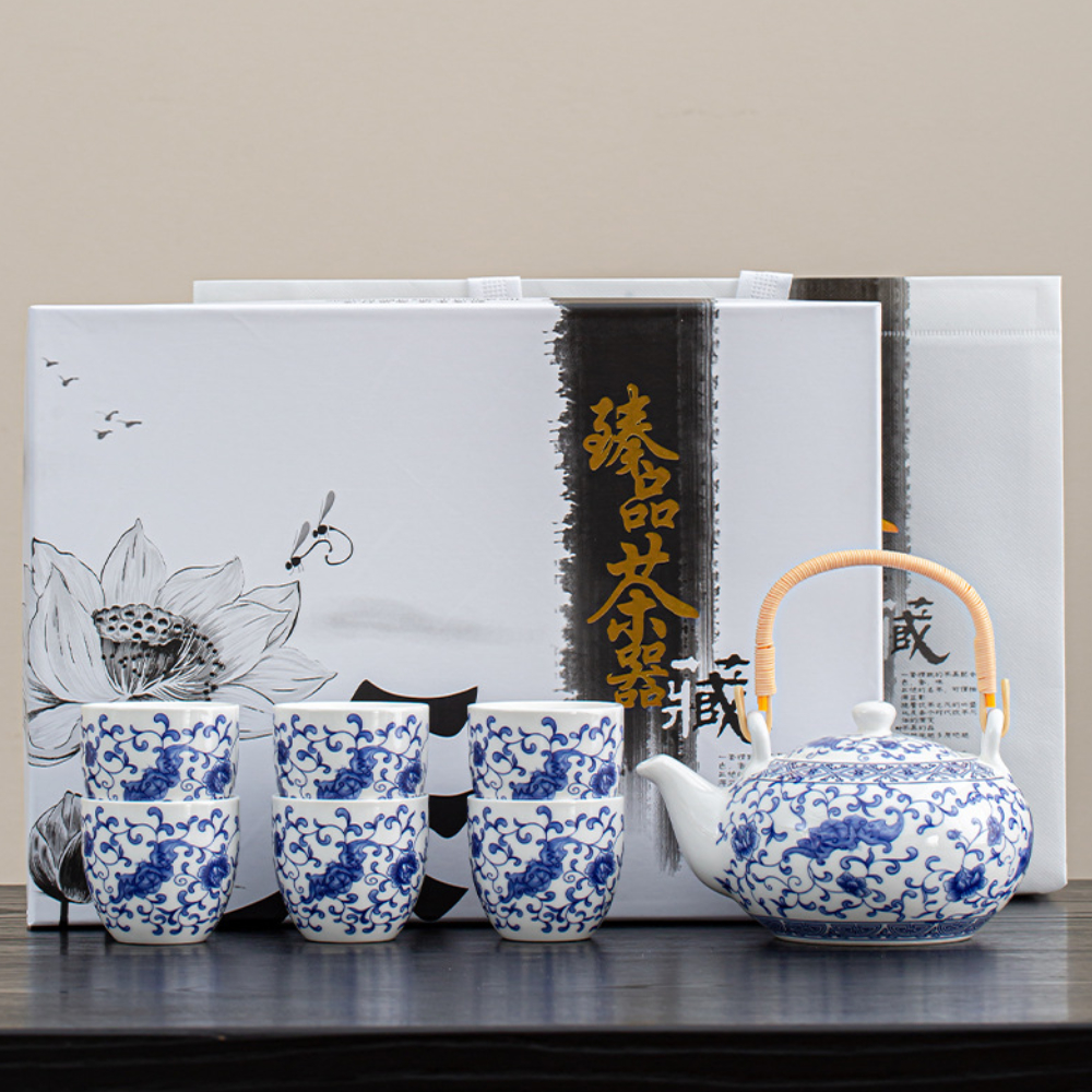 A blue and white porcelain tea set with a teapot and four cups is displayed in front of decorative packaging with floral and calligraphy designs.