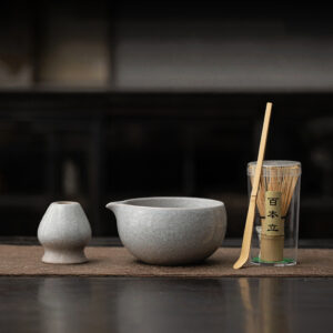 The Kiln-Glazed Matcha Set – 4 Piece | Stone includes a small cup, pouring bowl, bamboo whisk in a clear container, and bamboo scoop on a mat for an authentic tea experience.