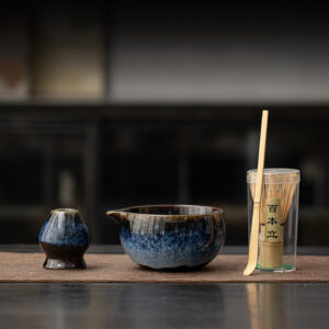 The Kiln-Glazed Matcha Set – 4 Piece | Stone, including a ceramic cup, spouted bowl, and bamboo whisk in a clear container, is displayed on a brown mat with a softly blurred stone-toned background.