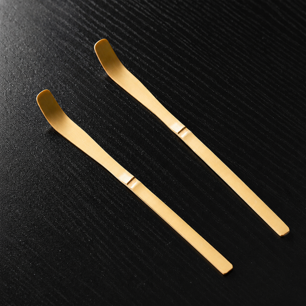 Two Bamboo Matcha Scoop - Chashaku, gold-colored metal spoons with flat, angled ends, lie parallel on a black textured surface.