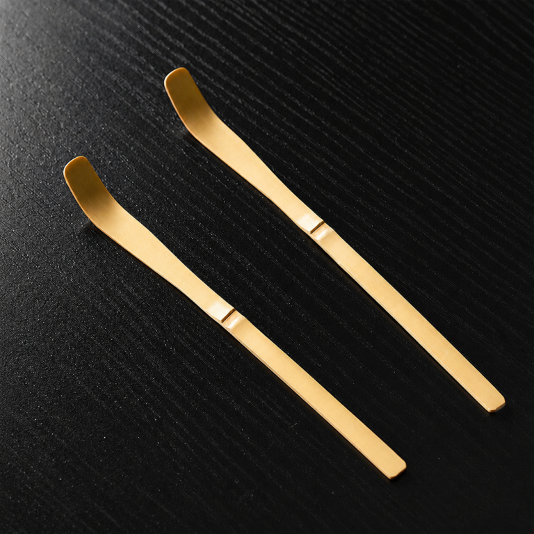 Two Bamboo Matcha Scoop - Chashaku, gold-colored metal spoons with flat, angled ends, lie parallel on a black textured surface.