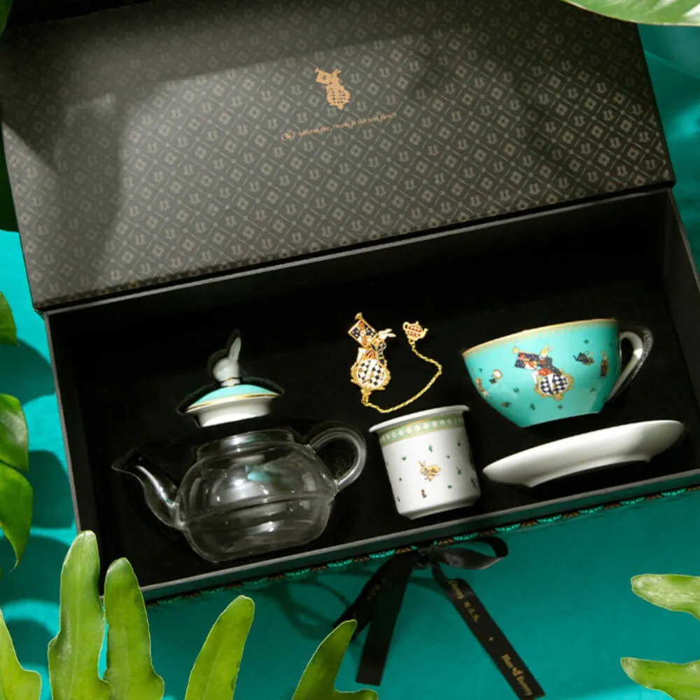 A decorative tea set in an open box with a patterned interior lid. Includes a glass teapot, a turquoise cup, a white cup, a saucer, and a golden accent piece. Surrounded by green leaves.