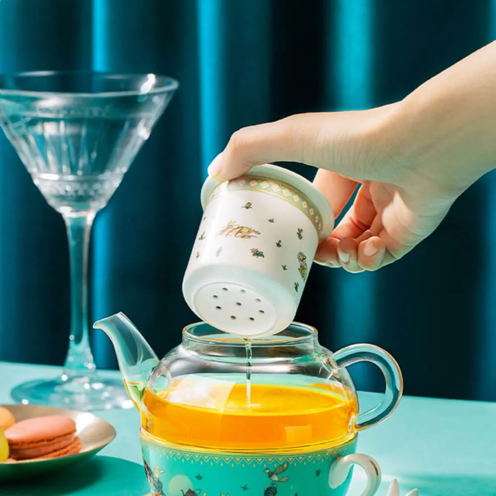 Hand pouring tea from a small strainer into a glass teapot, on a table with a martini glass and macarons.