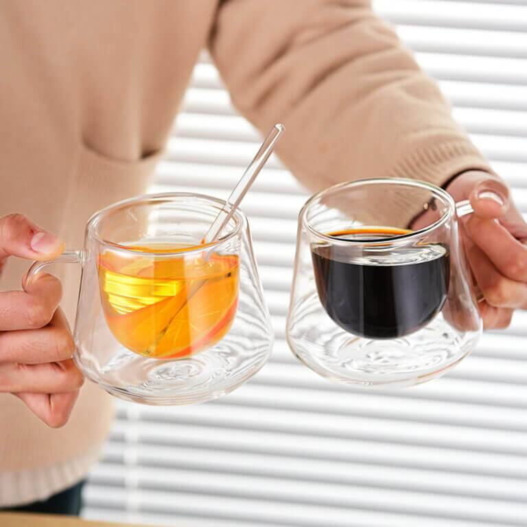 A person holds two Diamond Pattern Double-Wall Glass Cups—one with orange tea and a spoon, the other with black coffee—against white blinds. The insulated tumblers elegantly maintain the perfect temperature for each drink.
