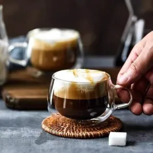 A hand reaches for a Double-Wall Glass Cup | Floral Tea & Coffee Tumbler (150ml) filled with coffee and frothy milk on a woven coaster. A similar tumbler sits in the background, with a sugar cube nearby.