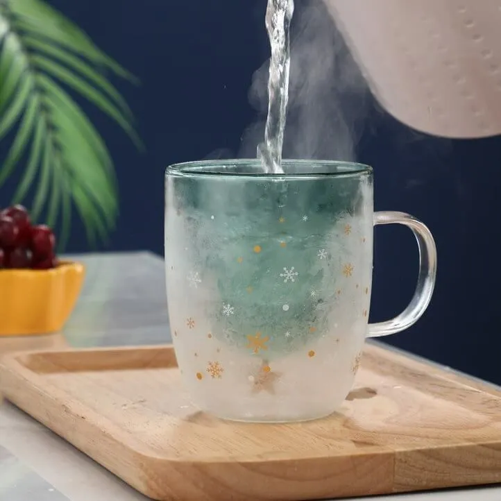 Hot water being poured into a frosted glass mug with snowflake motifs on a wooden tray. A bowl of grapes and a green plant are in the background.