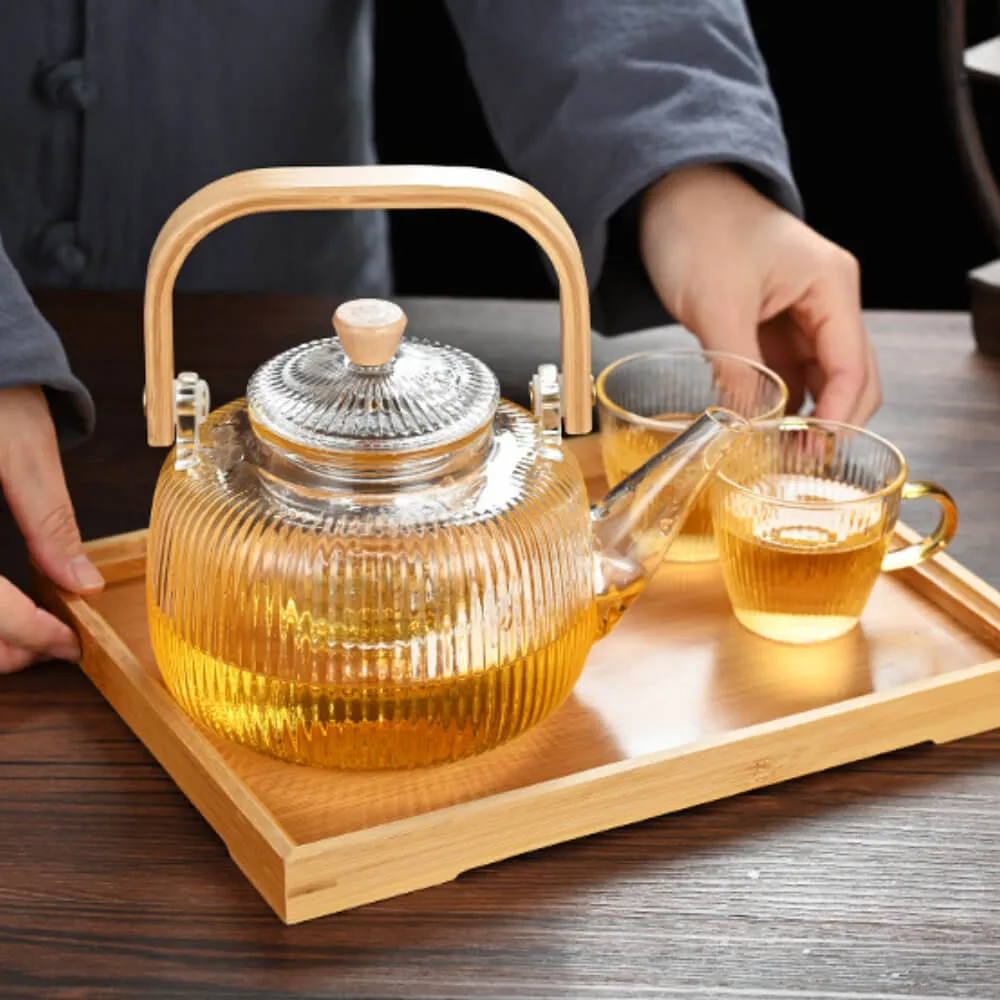 The Modern Glass & Bamboo Tea Set, featuring a sleek teapot and two matching teacups, sits elegantly on a wooden tray. A person carefully places the set on a wooden surface, setting the scene for a serene tea moment.