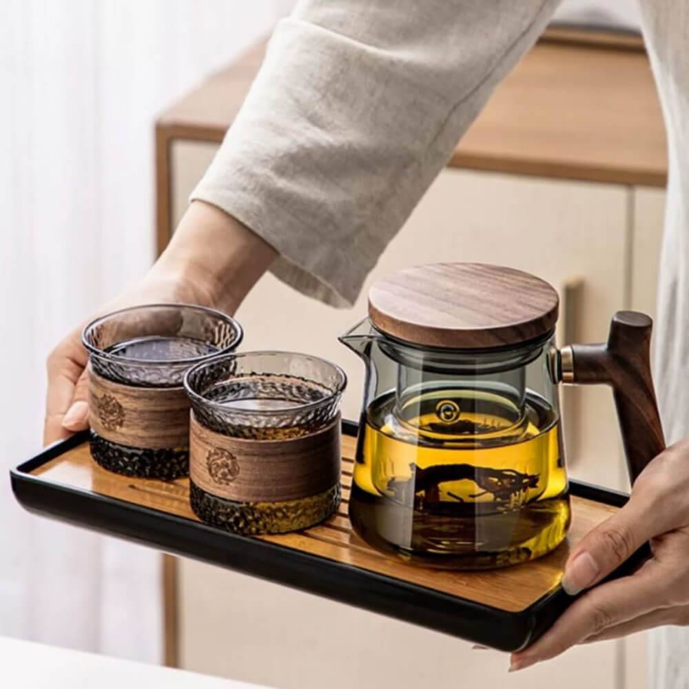 Someone showcases a Personal Glass Tea Master | Single-Serve Office Brewing Set, with a glass teapot containing yellow liquid and two glasses, all featuring wooden accents.