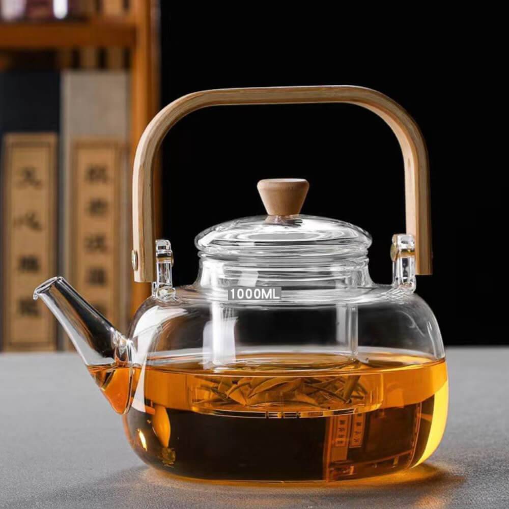 The Premium Glass Lifting Teapot, featuring a wooden handle and lid knob, is filled with amber Pu'er tea. The 1000ML heat-resistant kettle is set on a gray surface against a blurred background.