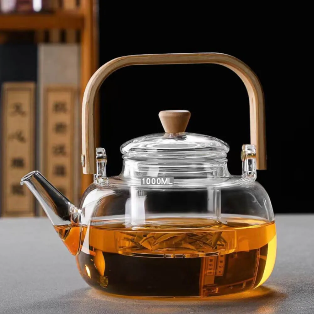 The Premium Glass Lifting Teapot, featuring a wooden handle and lid knob, is filled with amber Pu'er tea. The 1000ML heat-resistant kettle is set on a gray surface against a blurred background.