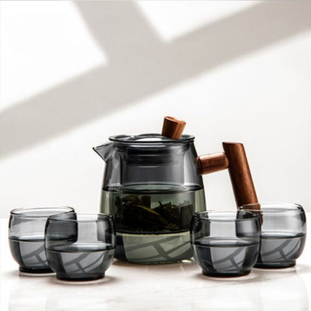 The Mountain View Glass Teapot, featuring a Japanese-style side handle with a wooden accent, sits on a surface beside four matching glass cups, each partially filled.