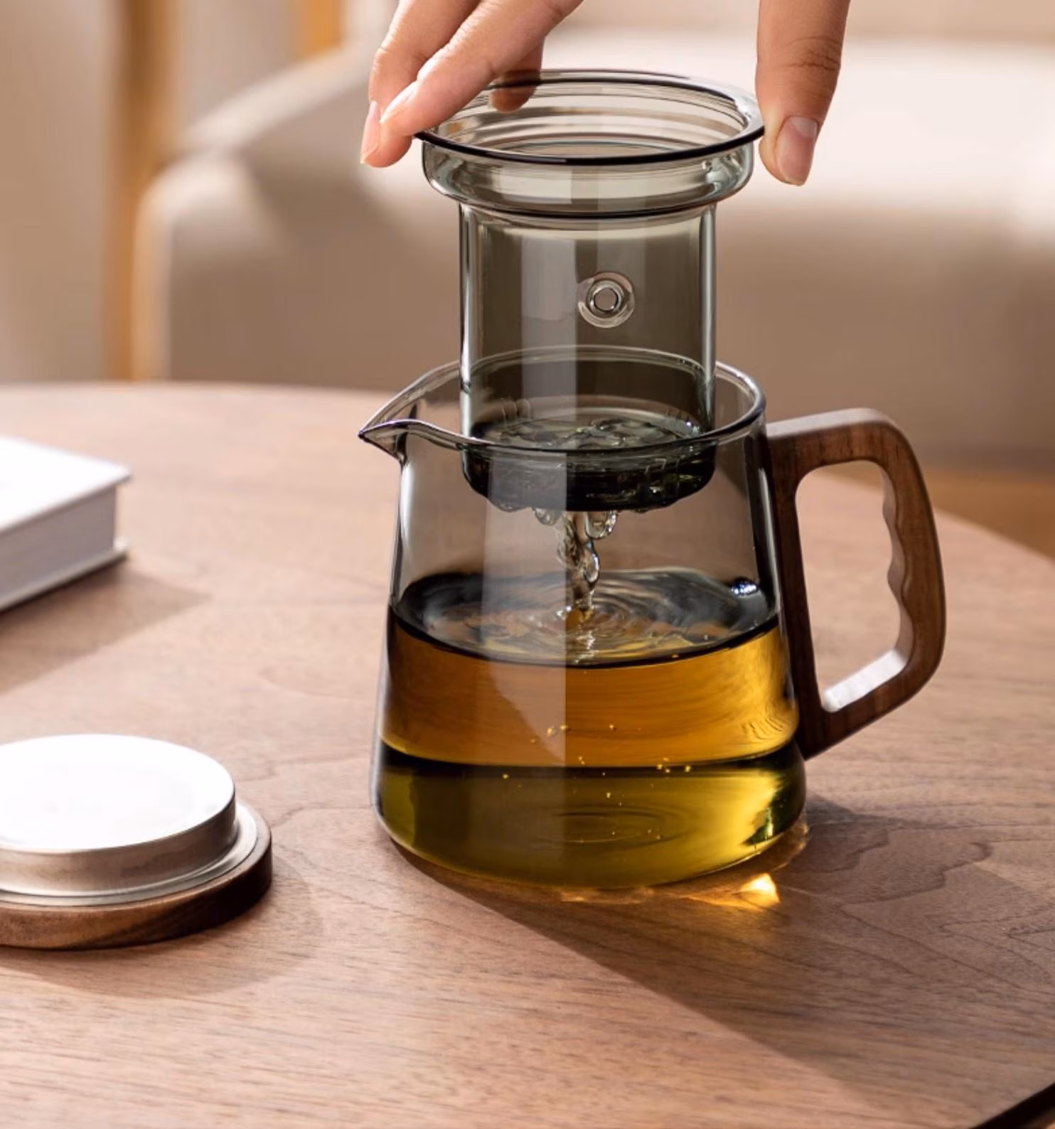 A hand places a glass infuser into a Borosilicate Glass Teapot with Glass Infuser & Walnut Handle filled with tea.